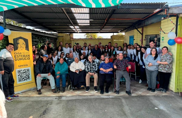 Fotografía grupal en la Casa Padre Esteban Gumucio. En un patio techado con láminas metálicas y una carpa a rayas turquesa, se reúnen aproximadamente 50 personas de diferentes edades. En primera fila, sentados, se encuentran varios adultos mayores, algunos en sillas de ruedas. De pie, detrás de ellos, hay un grupo numeroso de voluntarios principalmente jóvenes, muchos vistiendo camisetas blancas que parecen uniformes. El ambiente está decorado con globos de colores (azul, rojo y blanco) y guirnaldas. Las paredes del recinto son de diferentes colores: amarillo, verde limón y celeste.