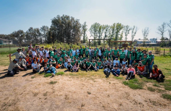 Grupo diverso de voluntarios de Zurich Santander con sus familias reunidos al aire libre para una foto grupal durante un proyecto comunitario ambiental. Participantes de diferentes edades con camisetas verdes, blancas y naranjas posando en un área verde con árboles. La escena representa una actividad colectiva llamada Reforestemos Juntos un Chile Nativo.