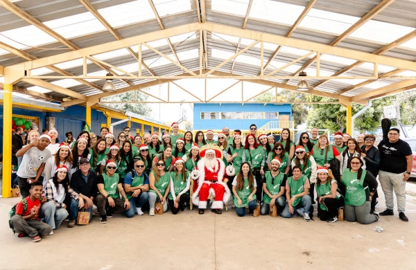 Fotografía de un evento navideño de voluntariado bajo un techado con estructura metálica y pilares amarillos. Un numeroso grupo de aproximadamente 60 voluntarios, identificables por sus camisetas verdes con el logo ZSChile, posan junto a una persona disfrazada de Santa Claus sentada en el centro. La mayoría de los voluntarios llevan gorros navideños rojos y están organizados en filas: algunos de pie, otros arrodillados o sentados en el suelo. Al fondo se observa una construcción azul y un patio escolar. Actividad benéfica navideña en un establecimiento educativo.