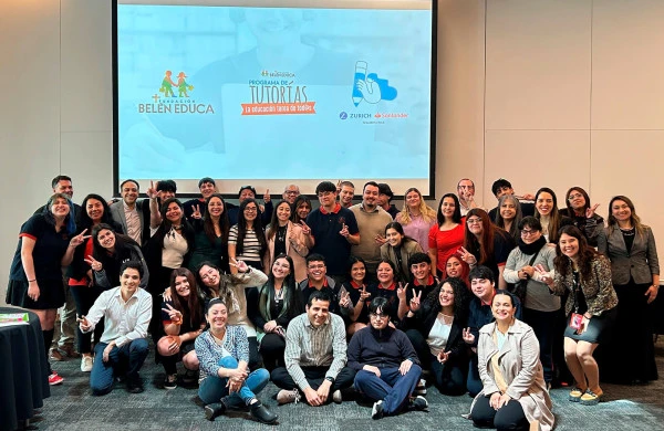 Fotografía grupal en un salón de conferencias, con aproximadamente 50 jóvenes adultos posando frente a una pantalla de proyección. En la pantalla se muestra el logo de Fundación Belén Educa y Programa de Tutorías con el lema La educación tarea de todos, junto con el logo de Zurich Santander. Los participantes están organizados en tres filas: algunos sentados en el suelo en primera fila, otros de pie, muchos haciendo el signo de la paz con los dedos. La imagen representa un encuentro de tutores o voluntarios educativos, posiblemente en una capacitación o cierre de programa. El ambiente es de celebración y compañerismo en un contexto educativo respaldado por empresas corporativas.