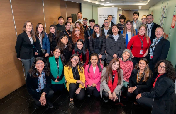 Fotografía grupal en un espacio corporativo moderno con paredes de madera y vidrio. Aproximadamente 40 jóvenes profesionales, principalmente mujeres, posan en varios niveles. En primera fila destacan personas con blazers coloridos en amarillo, rosa, verde y rojo. En el fondo se puede ver un banner blanco con el mensaje Juntos protegemos #LoQueMásImporta. El grupo forma parte de un evento o programa corporativo, todos visten de manera formal o semi-formal, y muchos llevan credenciales de identificación. La imagen transmite un ambiente profesional de networking o capacitación corporativa.