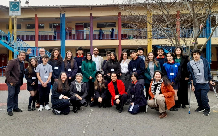 Fotografía grupal tomada en el patio de un colegio. En la imagen se observa a un grupo de aproximadamente 25 personas, entre estudiantes y adultos, posando y sonriendo frente a la cámara. Al fondo se aprecian columnas de colores y una estructura de dos pisos con pasillos y barandas azules.
