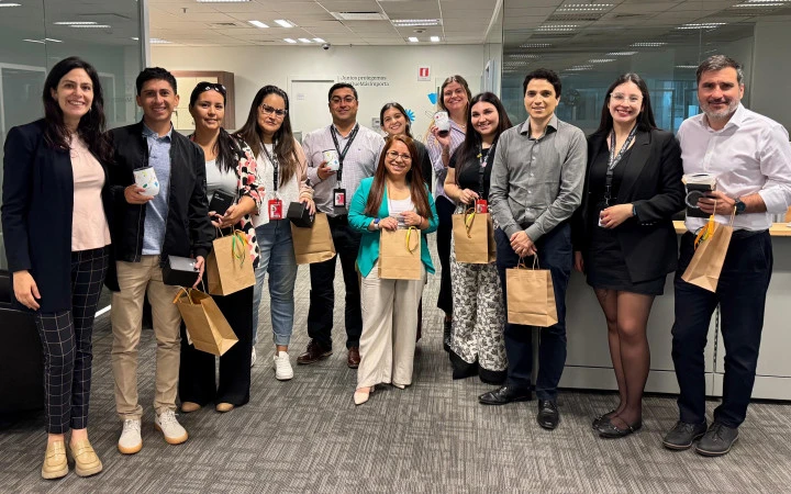Fotografía grupal tomada en un espacio de oficina. Aparecen alrededor de 13 personas adultas, mujeres y hombres, de pie y sonriendo a la cámara. Visten ropa formal y semiformal de trabajo. Varias de ellas sostienen bolsas de regalo de color café. El grupo se encuentra en un ambiente corporativo moderno, con escritorios y luz artificial. La imagen corresponde a un reconocimiento a voluntarios y voluntarias de Zurich Santander por su participación en actividades de voluntariado.