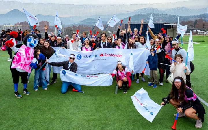 Fotografía grupal del equipo de Zurich Santander Chile durante la inauguración de las Olimpiadas del Seguro 2025. En primer plano, colaboradores y colaboradoras posan sonrientes al aire libre sosteniendo un lienzo de Zurich y Santander. A la izquierda aparece la mascota de la compañía, Zander. Detrás se observan banderas y globos, en un ambiente de celebración, compañerismo y entusiasmo deportivo.